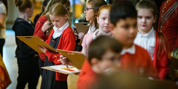 A group of school children in grey and red uniforms. Some of them are holding clipboards.