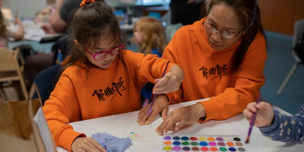 A mother and child, both wearing bright orange sweatshirts, are sitting at a table painting a picture together. In front of them is a box of paints.