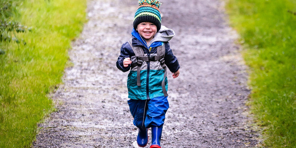 A small child dressed in a waterproof suit, wellington boots and a bobble hat is running along a path with grass on either side. The child is smiling.