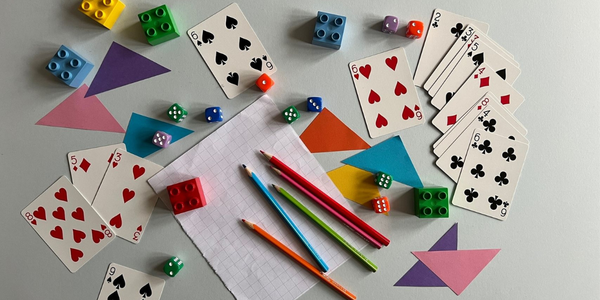 A photograph of maths activities props. In the middle is some note paper with coloured pencils. Around that is a spread out pack of cards, some dice, some coloured paper triangles, and some Lego Duplo blocks.