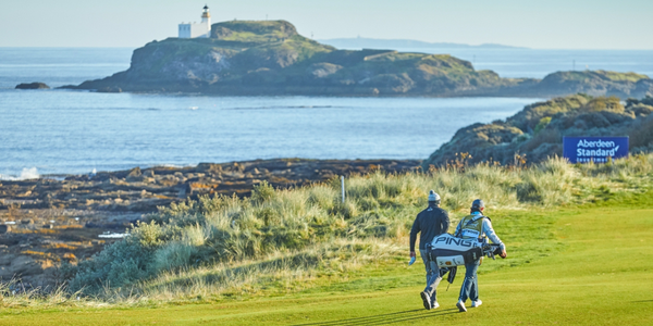 A photograph showing a section of a golf course. Two people are walking across the course, one of them is carrying a bag full of golf clubs. In the background you can see the sea, and an island with a lighthouse.