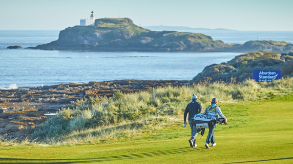 A photograph showing a section of a golf course. Two people are walking across the course, one of them is carrying a bag full of golf clubs. In the background you can see the sea, and an island with a lighthouse.