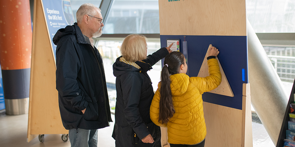 Two adults and a child are standing in front of a board that has "Why colour is maths?" written at the top. The child is adding a sticker to a triangle mounted on the board.